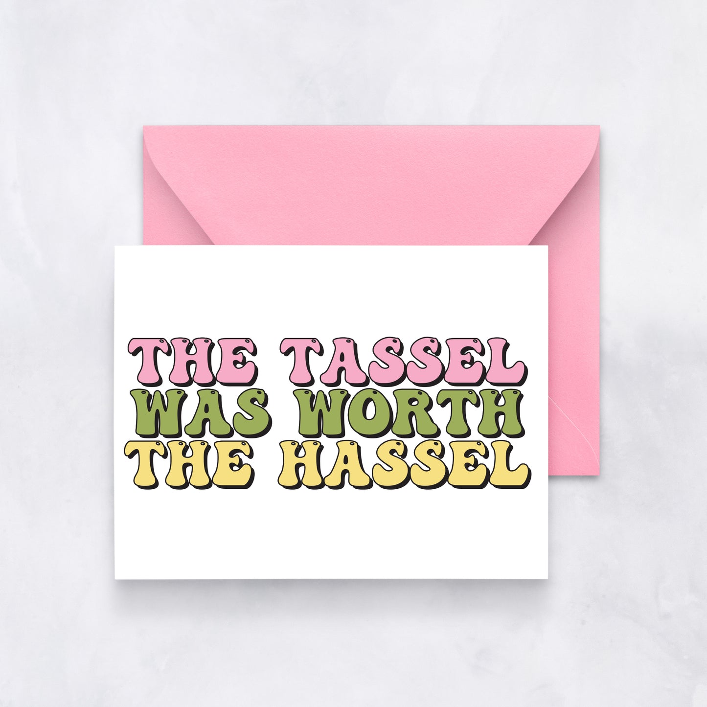 White card with bold retro text reading “The Tassel Was Worth the Hassel” in pink, green, and yellow, styled with a pink envelope.