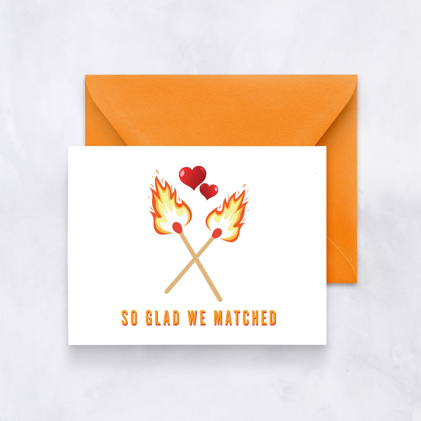 White card with two flaming matches crossed and hearts above, text reads “So glad we matched,” paired with an orange envelope.