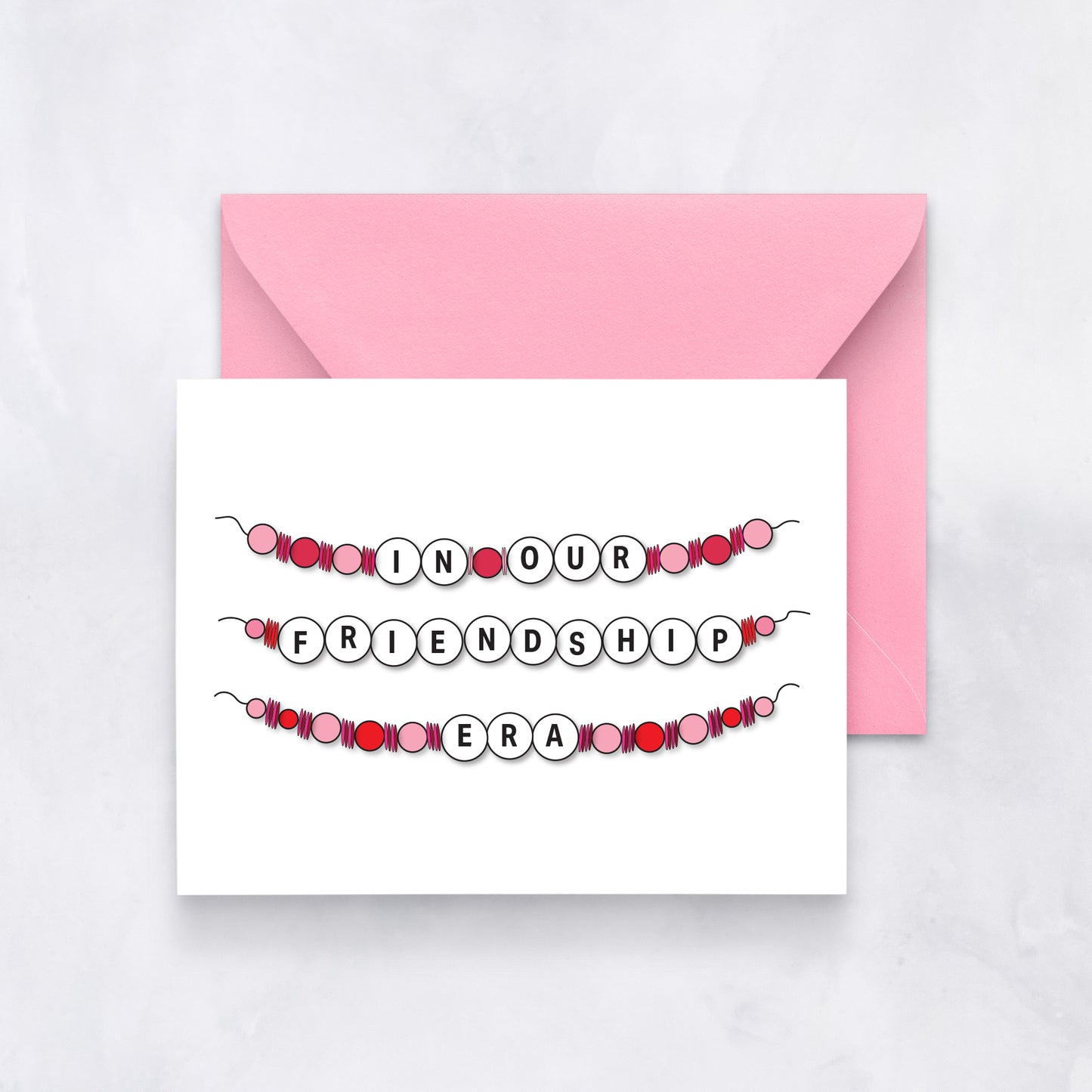 White card with beaded bracelet design spelling out “In Our Friendship Era,” paired with a pink envelope.