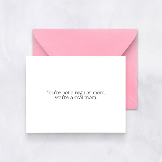 White greeting card with black text reading “You’re not a regular mom, you’re a cool mom,” styled with a pink envelope.