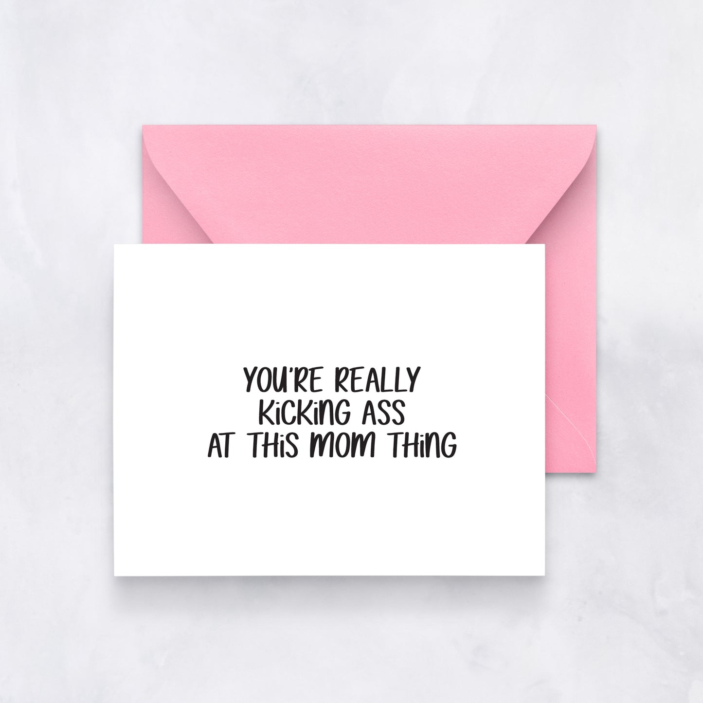 White greeting card with black handwritten text reading “You’re really kicking ass at this mom thing,” styled with a pink envelope.