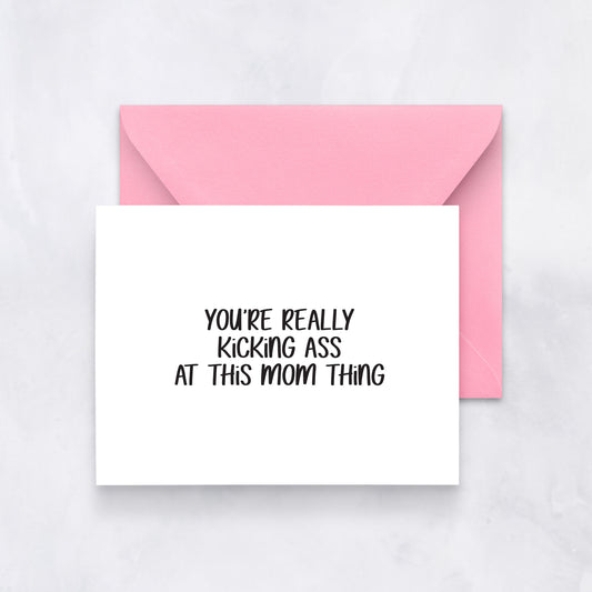 White greeting card with black handwritten text reading “You’re really kicking ass at this mom thing,” styled with a pink envelope.