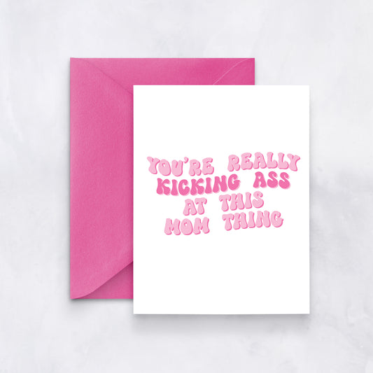 White greeting card with pink bubble text reading “You’re really kicking ass at this mom thing,” paired with a bright pink envelope.