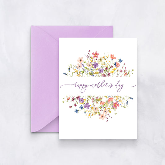 White greeting card with a watercolor wreath of colorful wildflowers and purple script reading “Happy Mother’s Day,” paired with a lavender envelope.