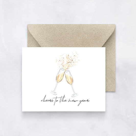 White greeting card with two watercolor champagne glasses clinking and text “cheers to the new year,” styled with a tan envelope.