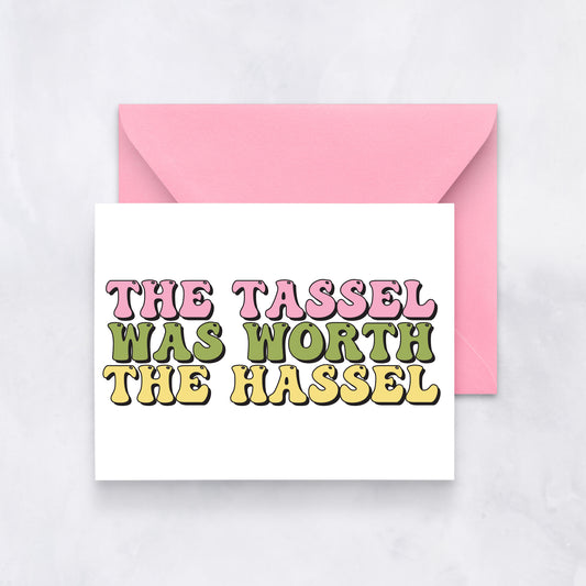 White card with bold retro text reading “The Tassel Was Worth the Hassel” in pink, green, and yellow, styled with a pink envelope.