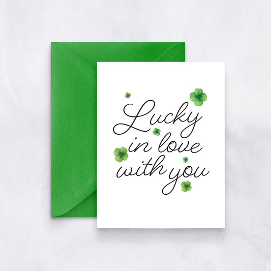 White card with black script reading “Lucky in love with you” surrounded by watercolor clovers, styled with a green envelope.