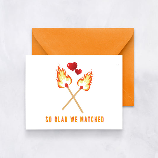 White card with two flaming matches crossed and hearts above, text reads “So glad we matched,” paired with an orange envelope.