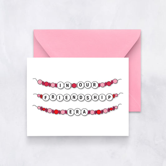 White card with beaded bracelet design spelling out “In Our Friendship Era,” paired with a pink envelope.