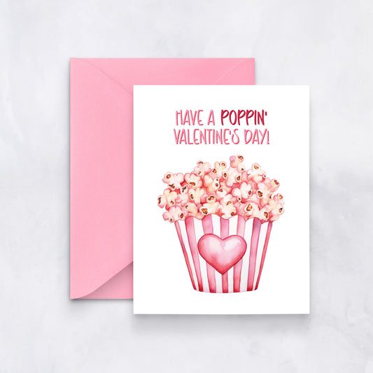 White card with pink watercolor popcorn bucket and heart design, text reads “Have a poppin’ Valentine’s Day,” styled with a pink envelope.