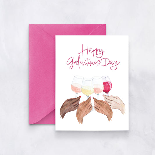 White card with watercolor hands holding wine glasses and pink text reading “Happy Galentine’s Day,” styled with a pink envelope.