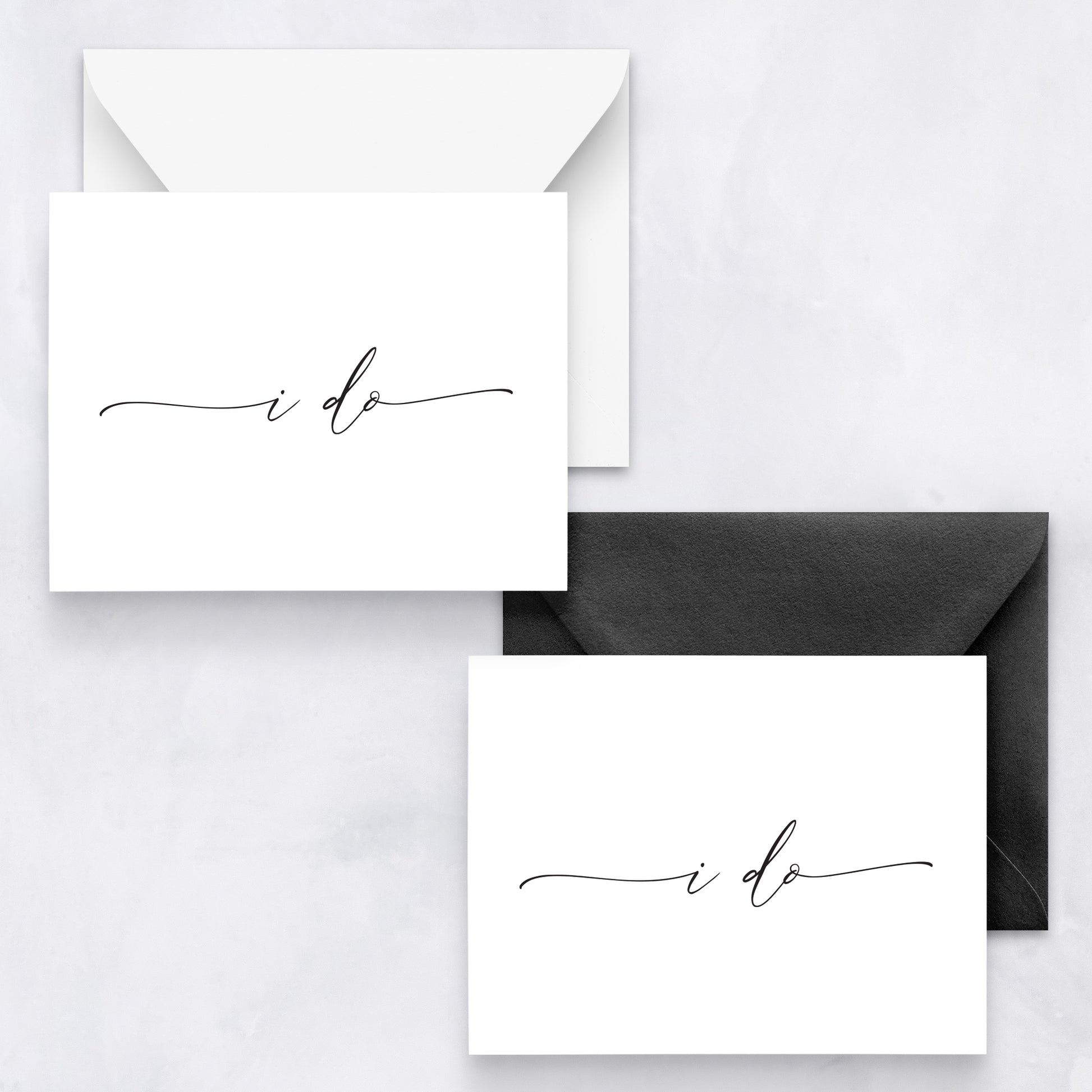 Two white wedding cards with black cursive text “i do,” styled with white and black envelopes on a marble background.