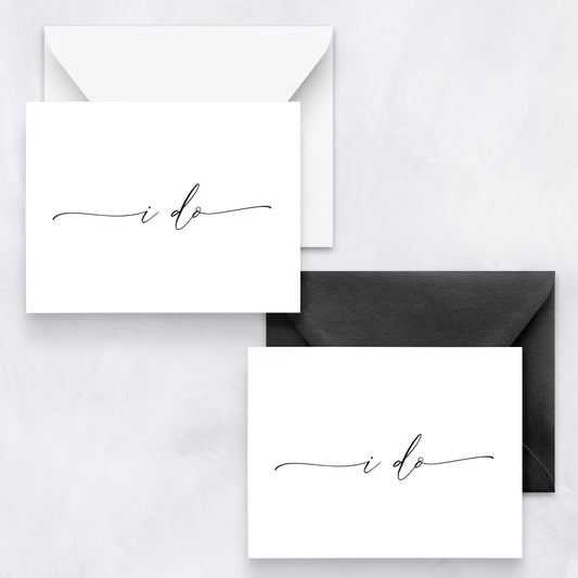 Two white wedding cards with black cursive text “i do,” styled with white and black envelopes on a marble background.