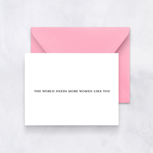 Minimal white card with small black serif text “THE WORLD NEEDS MORE WOMEN LIKE YOU,” paired with a light pink envelope.