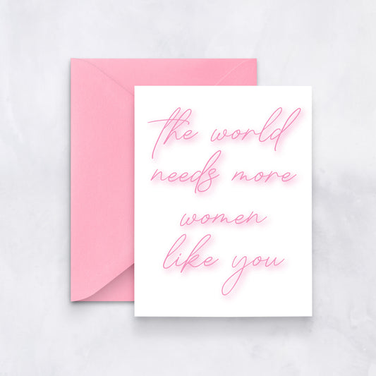 White card with neon-pink cursive text “the world needs more women like you,” paired with a soft pink envelope.
